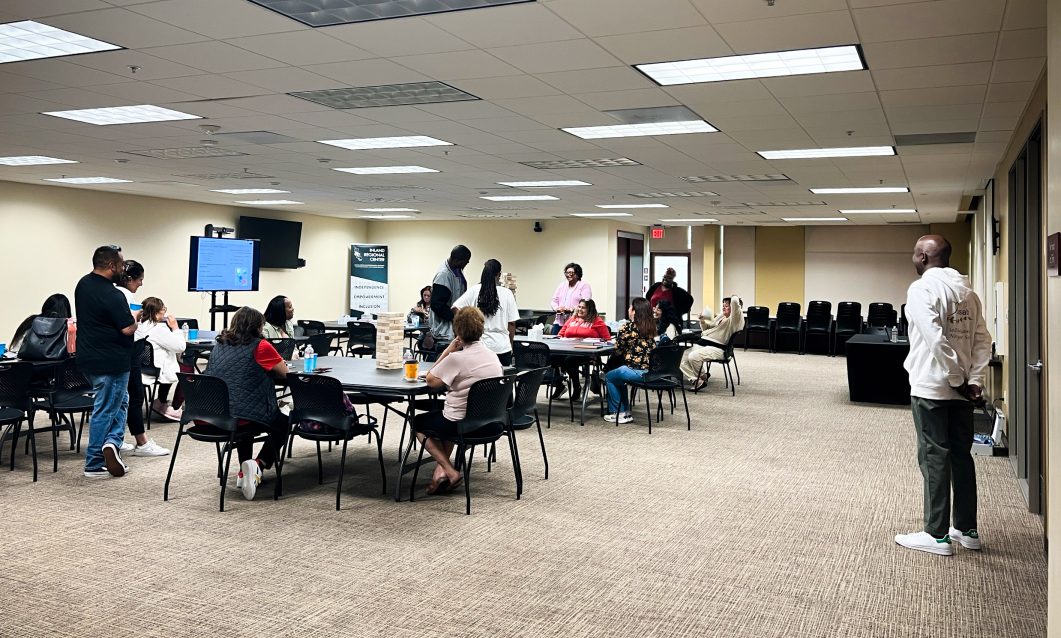 People sitting at tables in conference room, engaging in discussion, some standing near large screen.