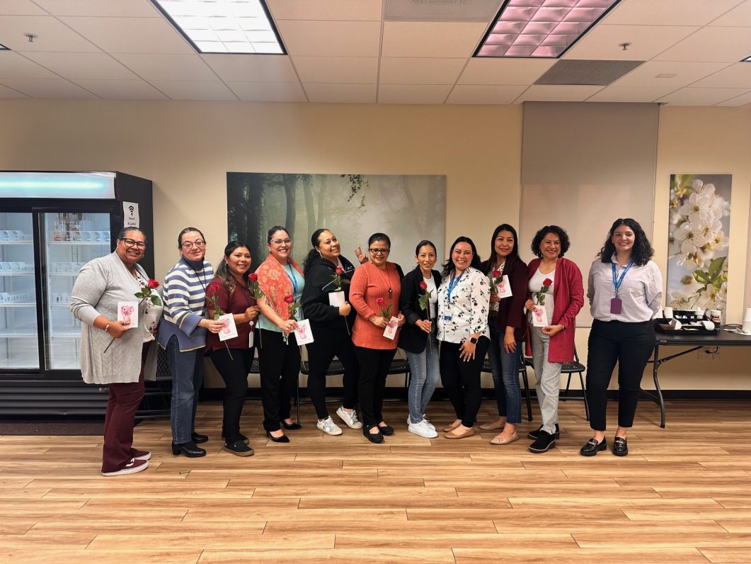 Group of eleven women standing, smiling, holding roses and cards in office setting.