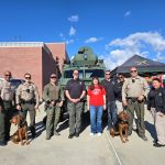 Group of people and two dogs in front of armored vehicle and tents under clear sky