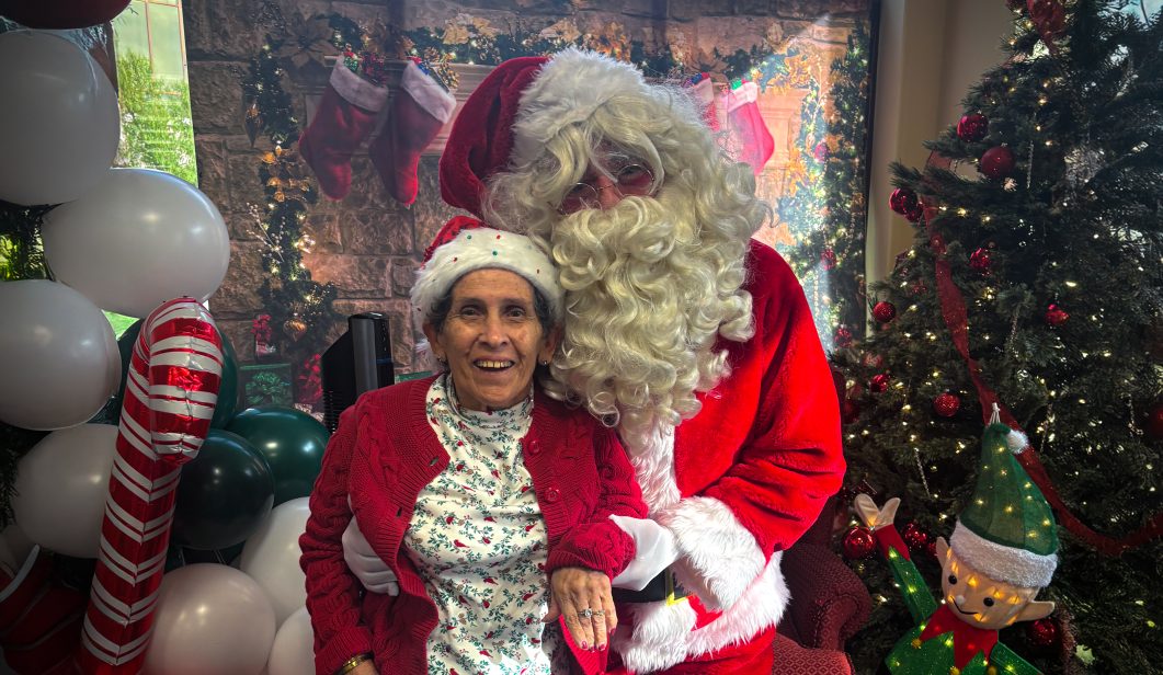 Elderly woman with Santa Claus in festive setting, surrounded by Christmas decorations and tree.