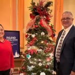Two people standing beside decorated Christmas tree with red and silver ornaments, illuminated lights.
