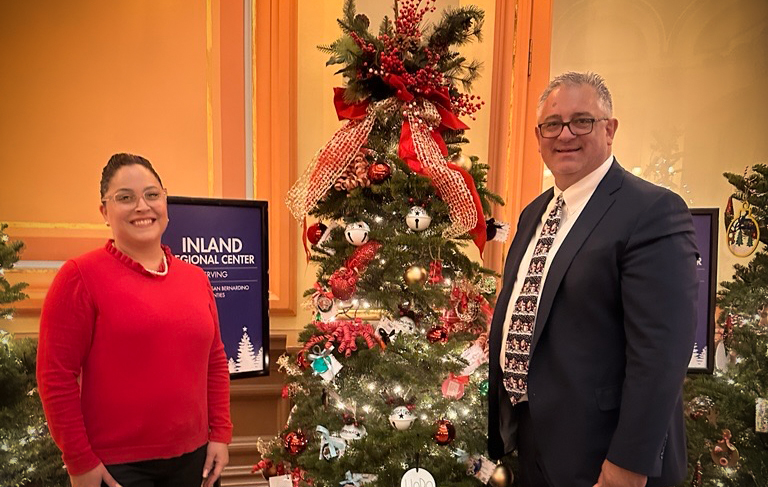 Two people standing beside decorated Christmas tree with red and silver ornaments, illuminated lights.
