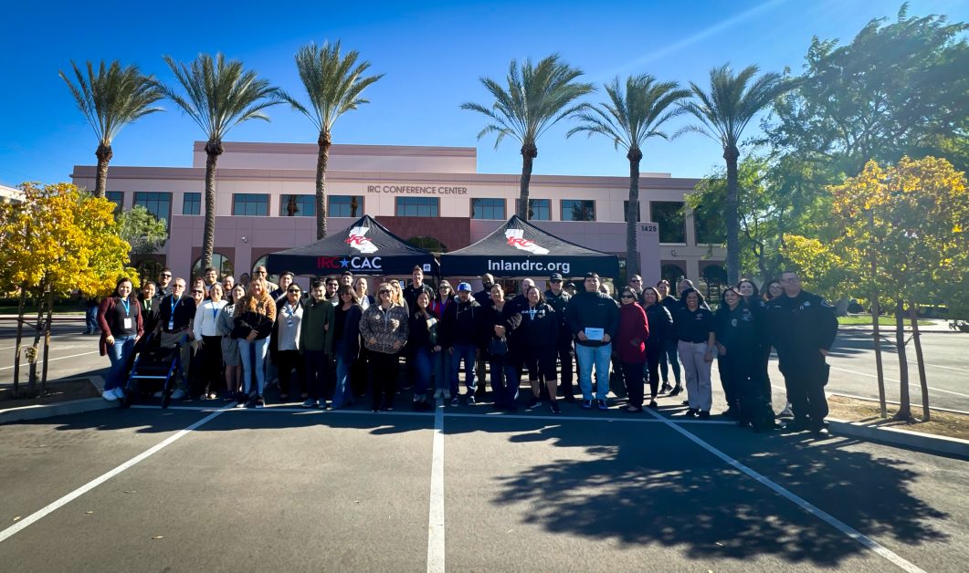 Group of people standing in parking lot beneath two black tents, palm trees in background.