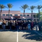 Group of people standing in parking lot beneath two black tents, palm trees in background.