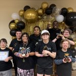 Eight people holding certificates and trophies, standing in front of gold and black balloon arch.