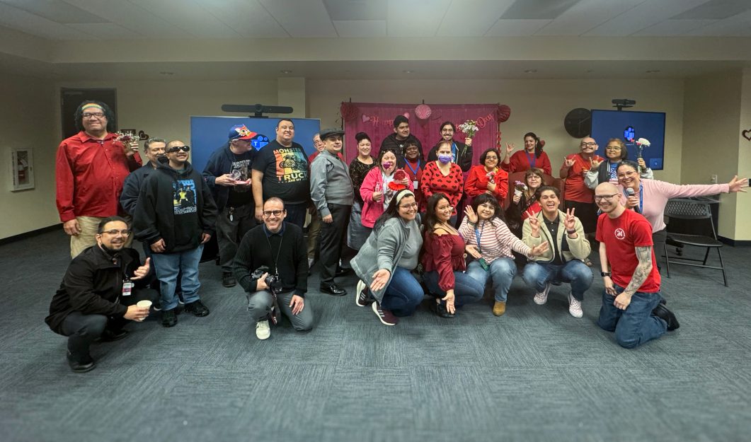 Group of people smiling indoors wearing red and black, holding flowers and making peace signs.