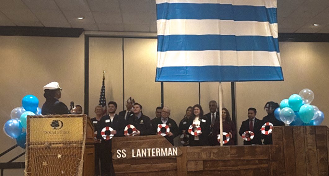 Group of people standing on stage with ship-themed decorations and blue-striped flag.