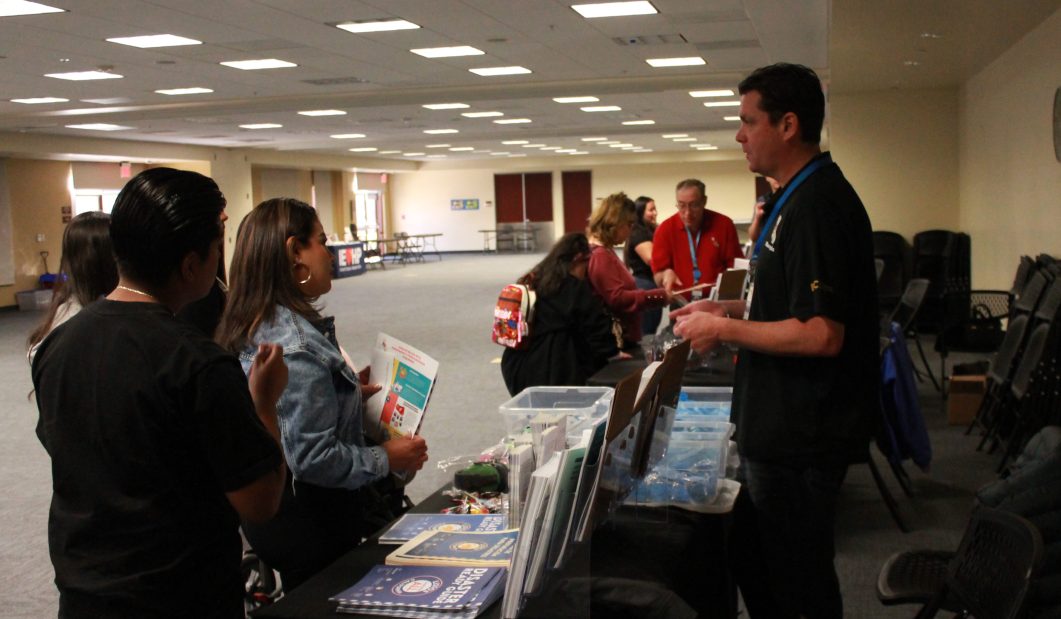 People attending information booth in large conference room, holding pamphlets and talking to exhibitors.
