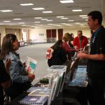 People attending information booth in large conference room, holding pamphlets and talking to exhibitors.