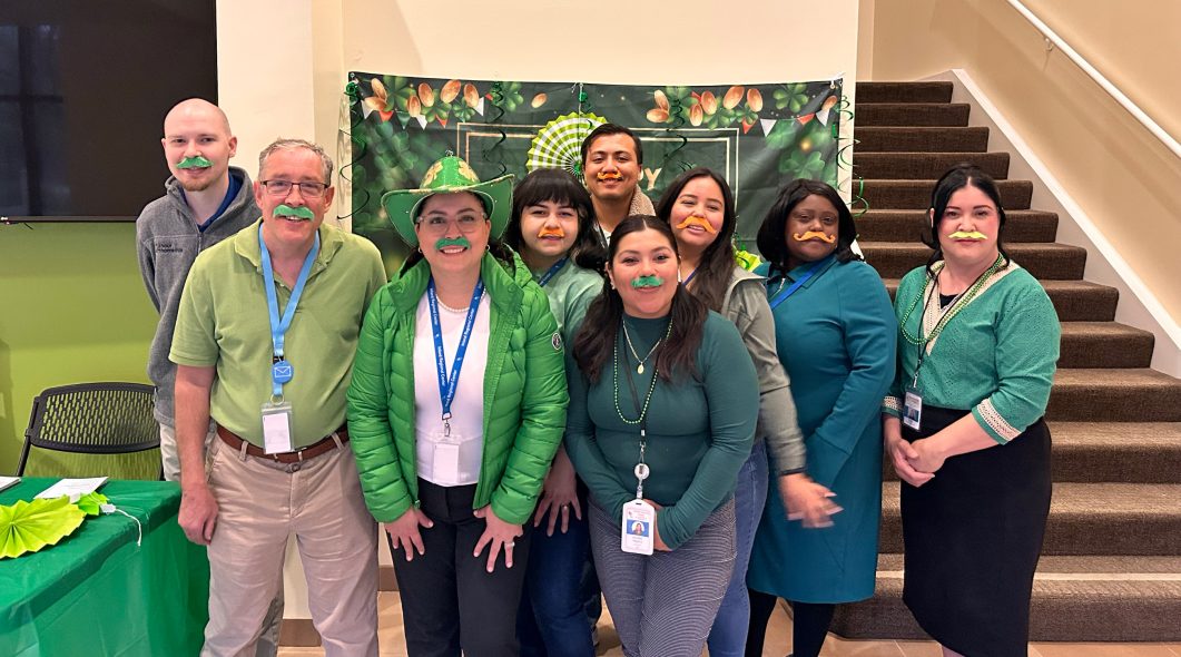 Group of people wearing green clothing and fake green mustaches, standing together smiling indoors.