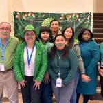 Group of people wearing green clothing and fake green mustaches, standing together smiling indoors.