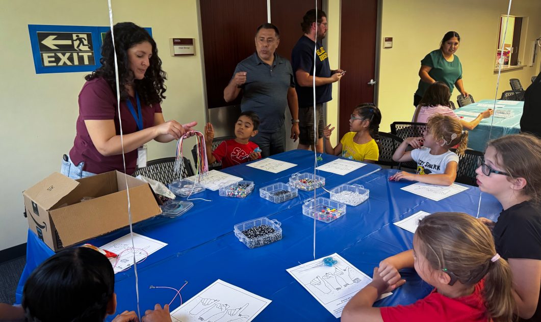 Children and adult engaged in crafting activity at table with colorful beads and strings.