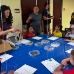 Children and adult engaged in crafting activity at table with colorful beads and strings.