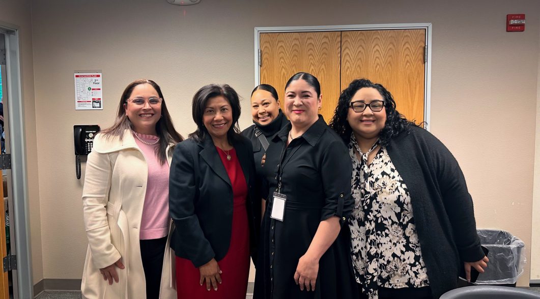 Five women standing together in office setting, smiling at camera, wearing business casual attire.