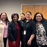 Five women standing together in office setting, smiling at camera, wearing business casual attire.