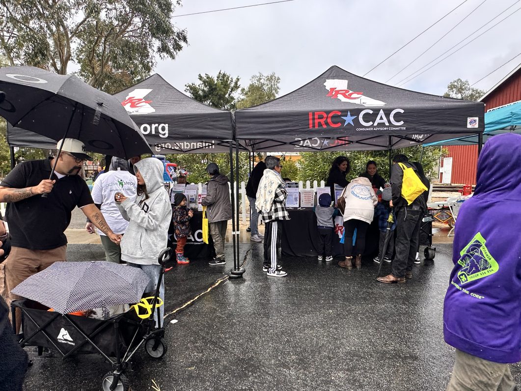 People gathering under black tents during rainy outdoor event, some holding umbrellas, colorful jackets visible.