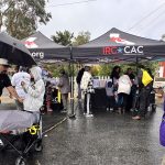 People gathering under black tents during rainy outdoor event, some holding umbrellas, colorful jackets visible.