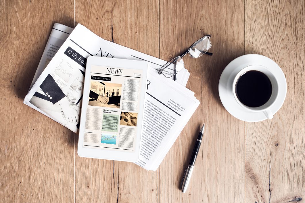 Tablet displaying news, stacked newspapers, eyeglasses, coffee cup, and pen on wooden surface.