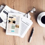 Tablet displaying news, stacked newspapers, eyeglasses, coffee cup, and pen on wooden surface.