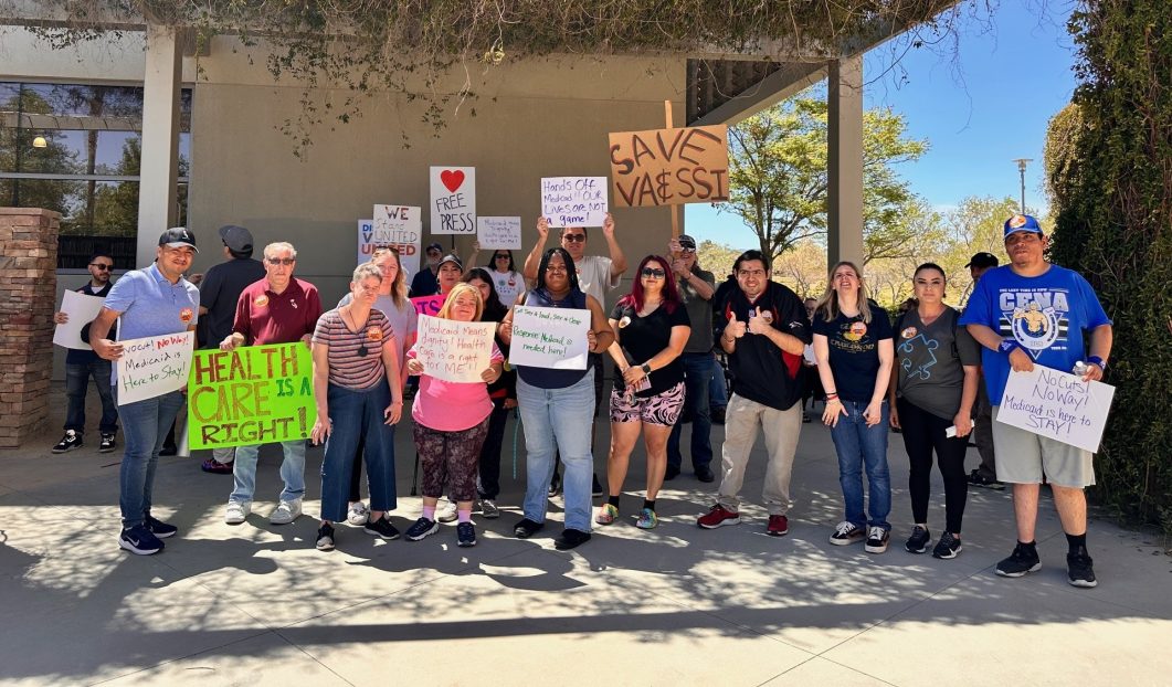 Group of people holding signs advocating for healthcare and Medicaid support outside building.