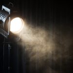Black stage spotlight emitting bright beam with visible mist or smoke in dark setting.