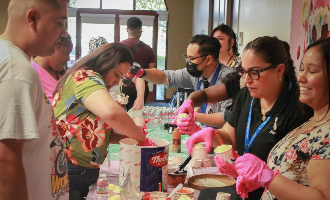 People serving ice cream at a table with cups, toppings, and colorful decorations.