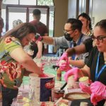 People serving ice cream at a table with cups, toppings, and colorful decorations.