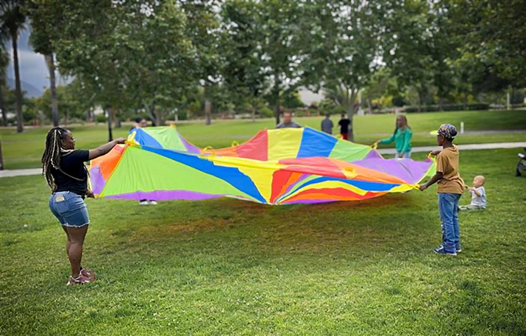 People holding colorful parachute in park, green grass, trees in background, baby sitting nearby.