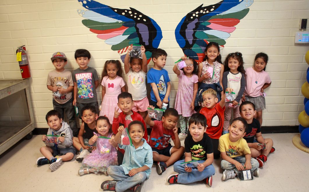 Group of smiling children in front of colorful painted wings on white brick wall.