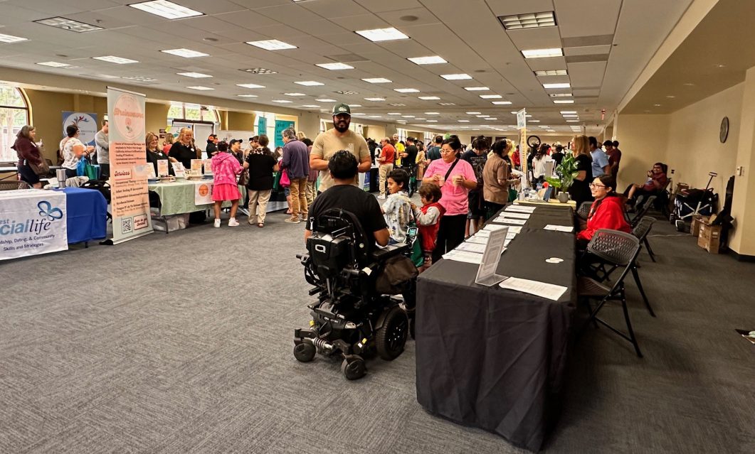 Crowded conference room with booths, people interacting, and person in wheelchair in foreground.