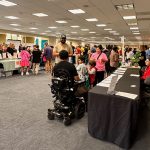 Crowded conference room with booths, people interacting, and person in wheelchair in foreground.