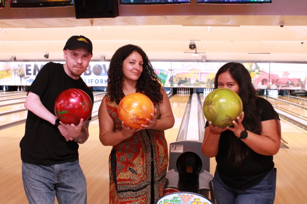 Three people holding colorful bowling balls at a bowling alley, smiling confidently at camera.