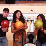 Three people holding colorful bowling balls at a bowling alley, smiling confidently at camera.