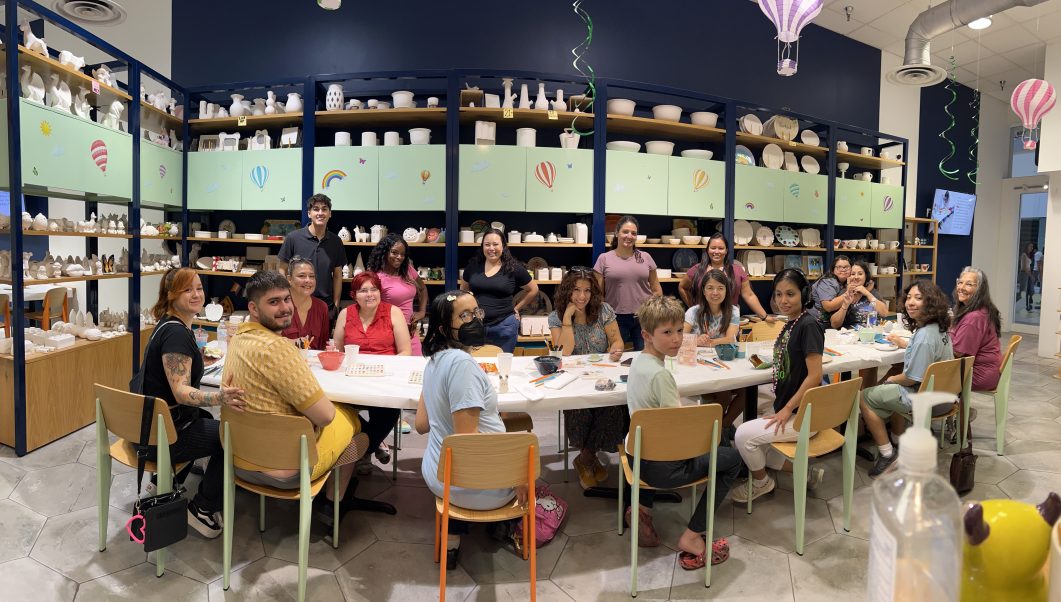 Group of people sitting around table in pottery studio with shelves of ceramics in background.