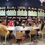 Group of people sitting around table in pottery studio with shelves of ceramics in background.