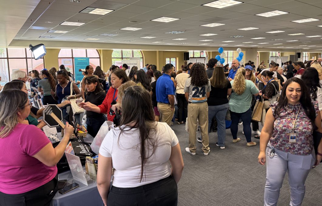 Crowded room with people interacting at tables, exchanging information, and holding tote bags.