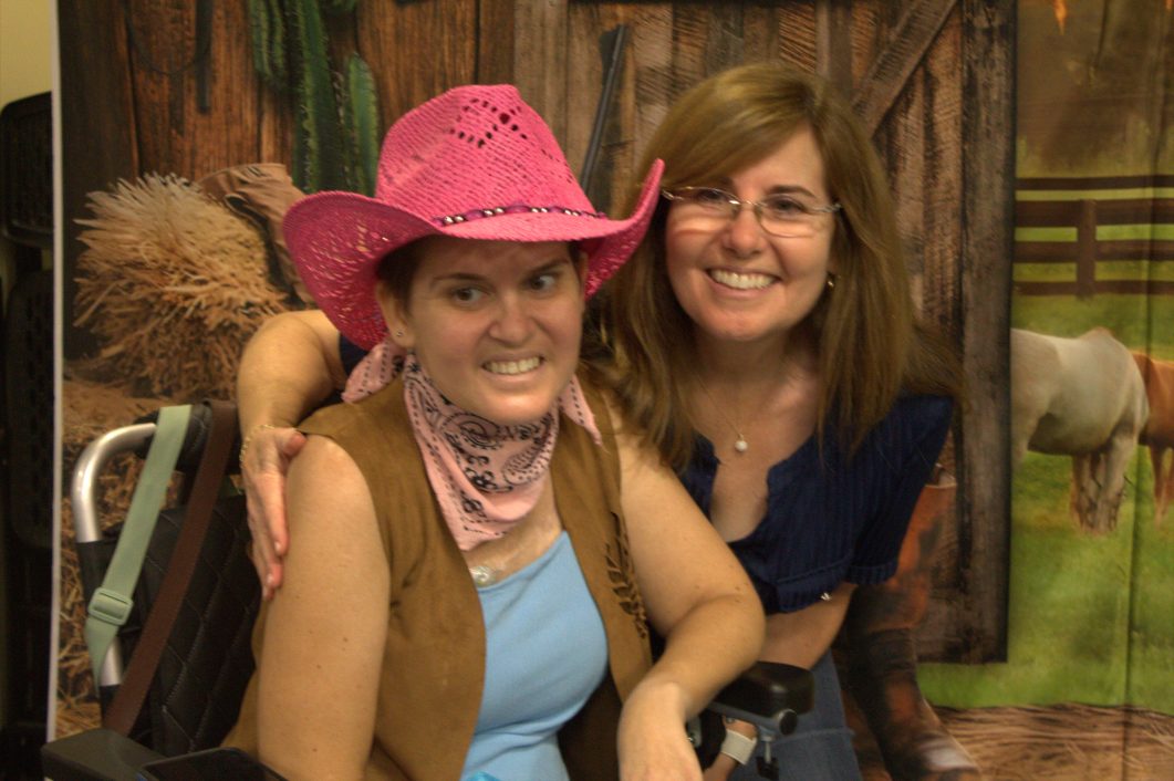Two women smiling, one in pink cowboy hat and scarf, in front of cowboy-themed backdrop.