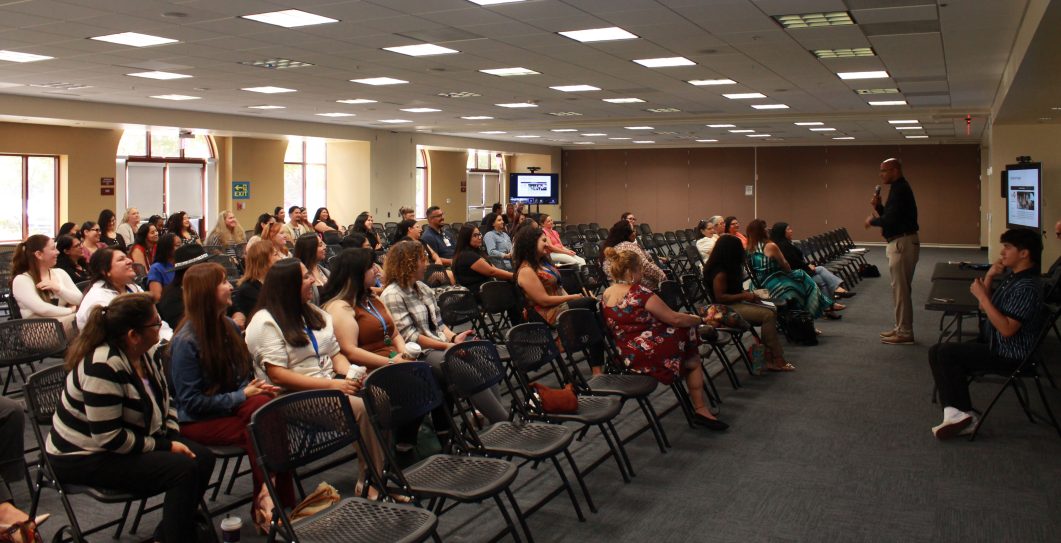 People seated attentively in conference room, speaker standing with microphone, presentation screen in background.
