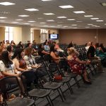 People seated attentively in conference room, speaker standing with microphone, presentation screen in background.