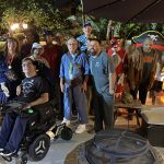 Group of people in casual clothes and costumes outdoors at night, with Halloween decorations.