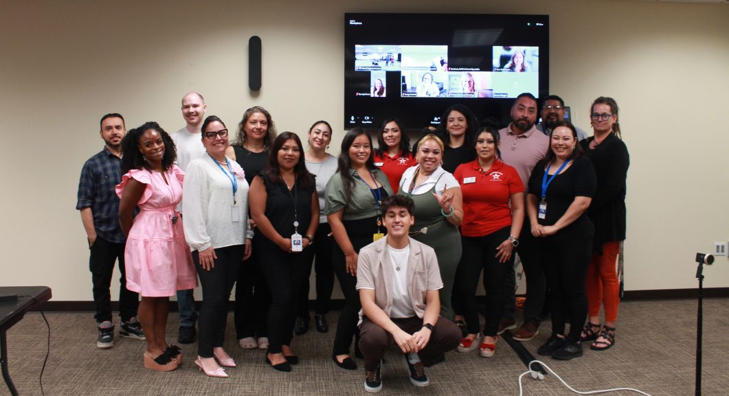 Group of cheerful adults posing indoors with video call screen in background.