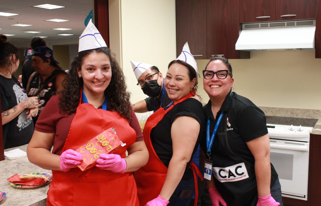 Four smiling people wearing paper hats and aprons, holding hot dog bag in kitchen.