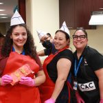 Four smiling people wearing paper hats and aprons, holding hot dog bag in kitchen.