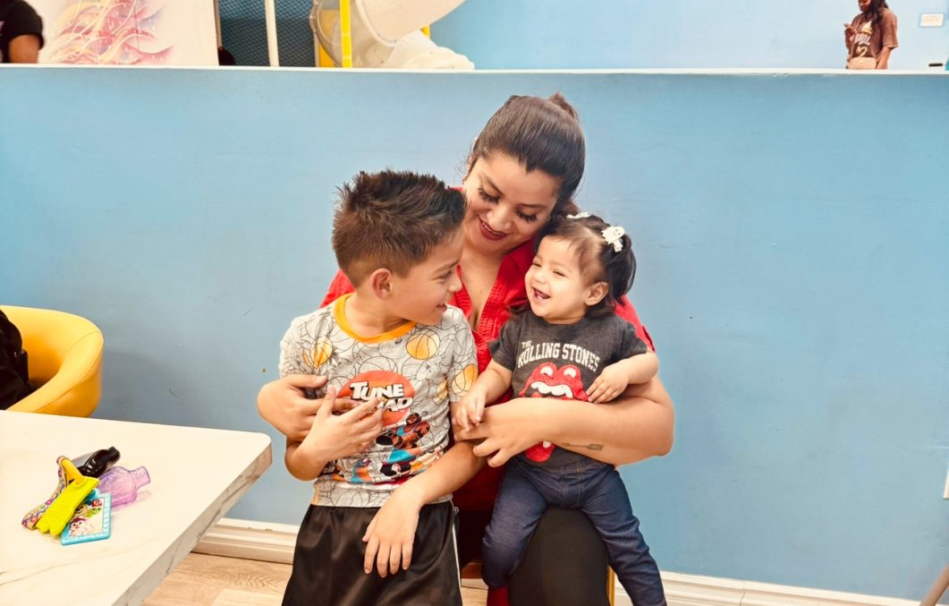 Woman in red shirt with two smiling children against blue wall indoor setting.