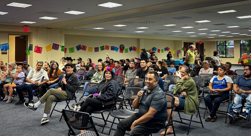 People seated in community room with colorful papel picado decorations, two holding microphones, some smiling.