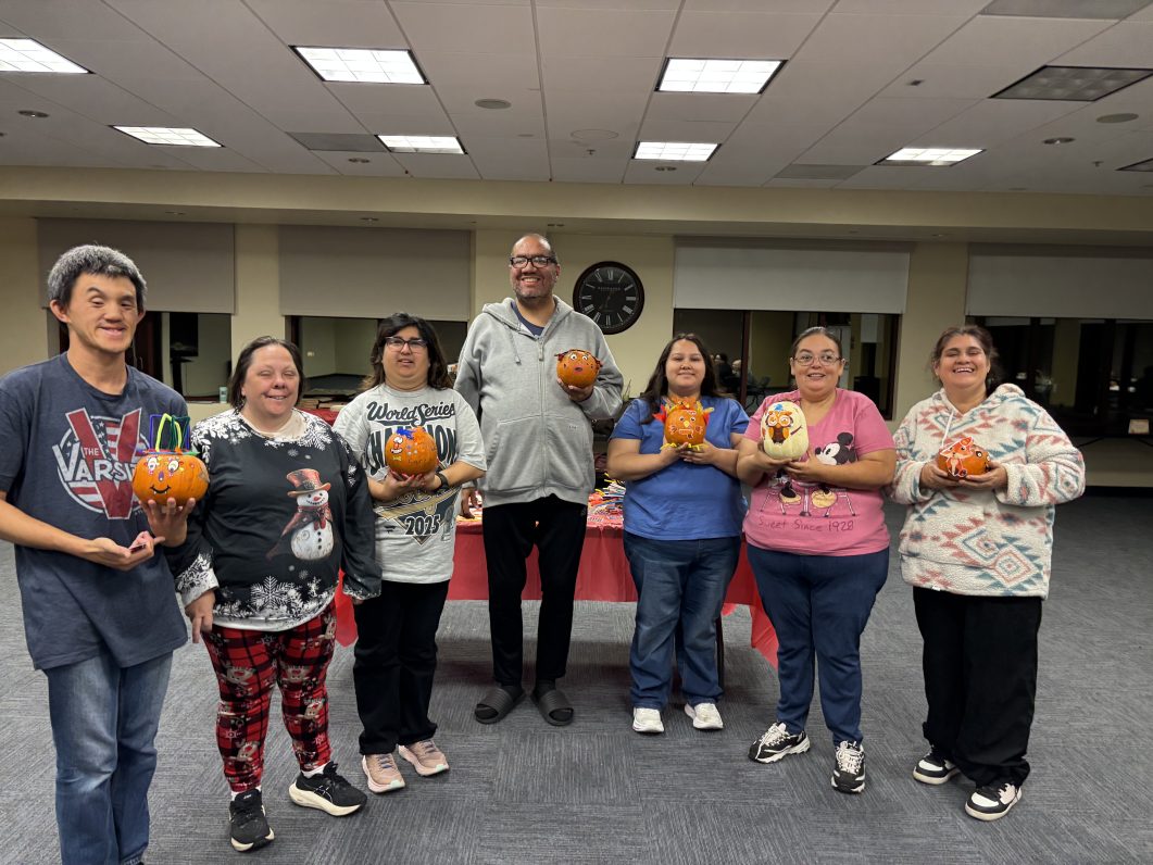 Group of seven people holding small decorated pumpkins inside brightly lit room.