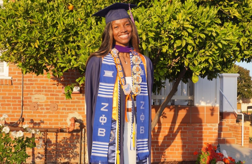 Graduating student wearing cap, gown, and sash stands in front of brick building and green tree.