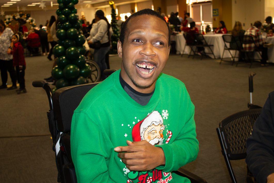 Smiling person in green holiday sweater, sitting in wheelchair at festive indoor gathering.