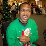 Smiling person in green holiday sweater, sitting in wheelchair at festive indoor gathering.
