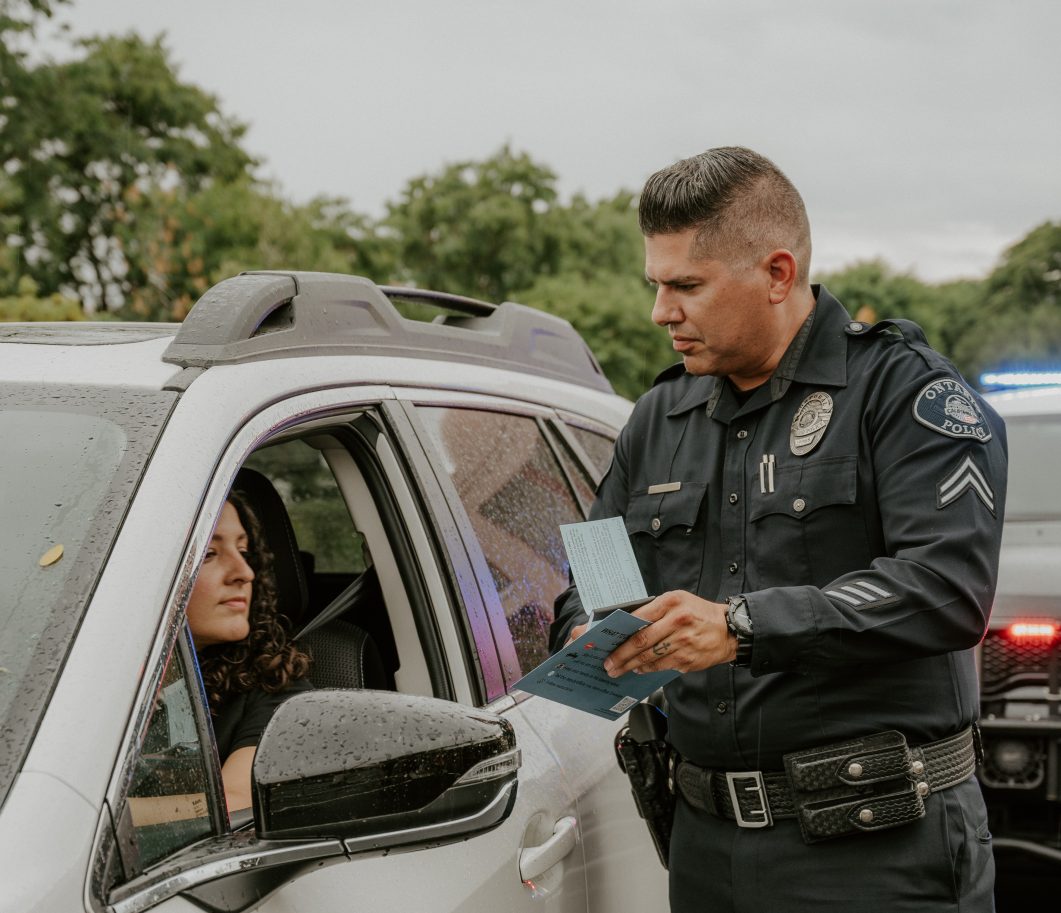Police officer standing next to car, talking to driver through open window, holding clipboard.